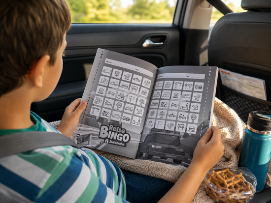 Kind sitzt auf der Rückbank und hält ein geöffnetes ReiseBingo Autobahn Buch während einer Autofahrt